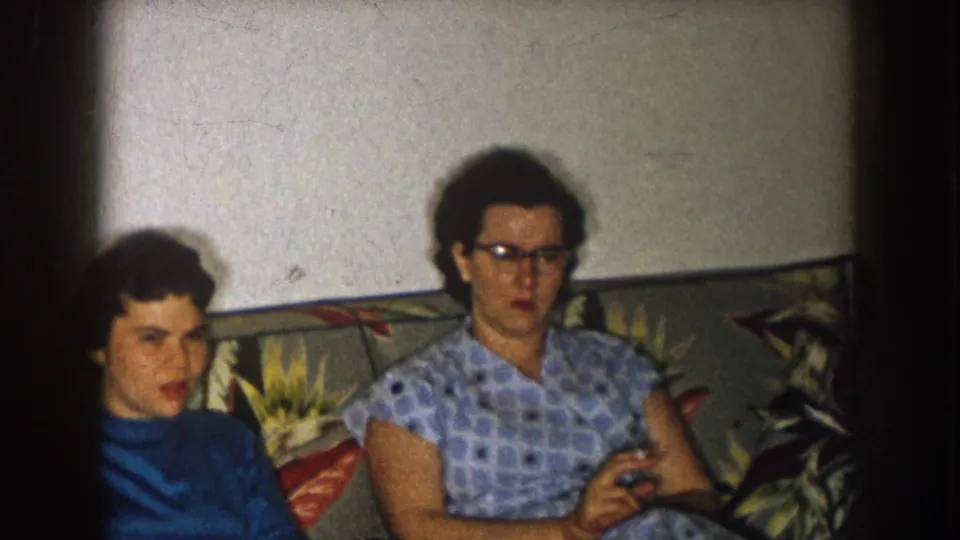 Two women remain seated on the floral sofa in the 1957 Nebraska home-movie scene, with one woman wearing glasses.
