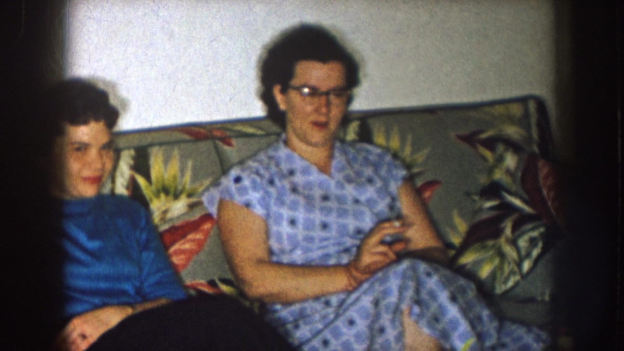Two women sit on a floral sofa, one in a blue turtleneck and one in a checked dress with glasses.