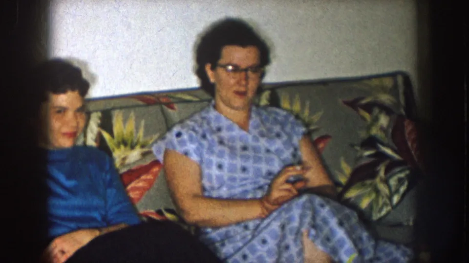 In a 1957 Nebraska home-movie frame, two women sit on a floral sofa indoors, with one holding a cigarette and the other wearing glasses.
