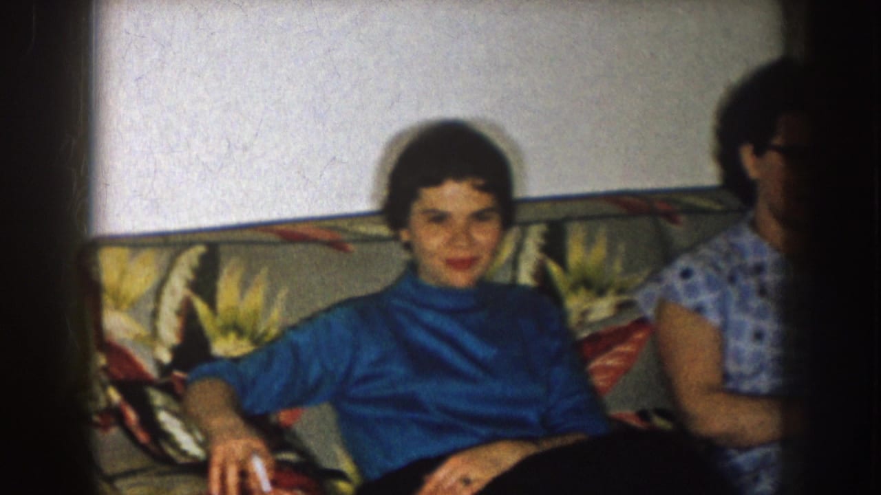 Woman in a blue turtleneck sits on a floral sofa while another woman is partly visible beside her.