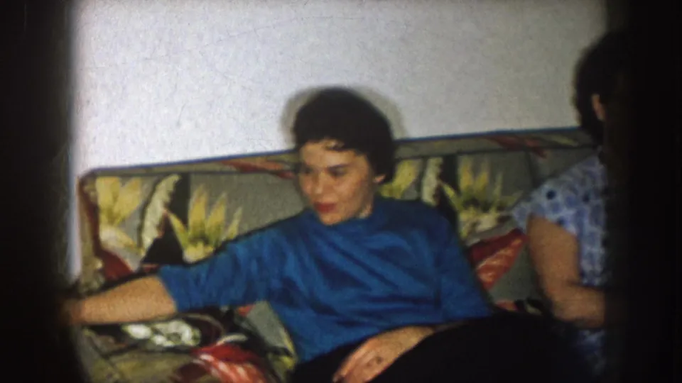 A seated woman in a blue turtleneck reaches toward the sofa arm in this 1957 Nebraska home-movie interior, with another woman partly visible.