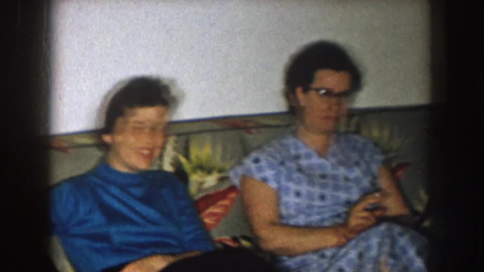 Two women sit on a floral sofa in a Nebraska home interior, one blurred in motion and the other wearing glasses and a blue checked dress.