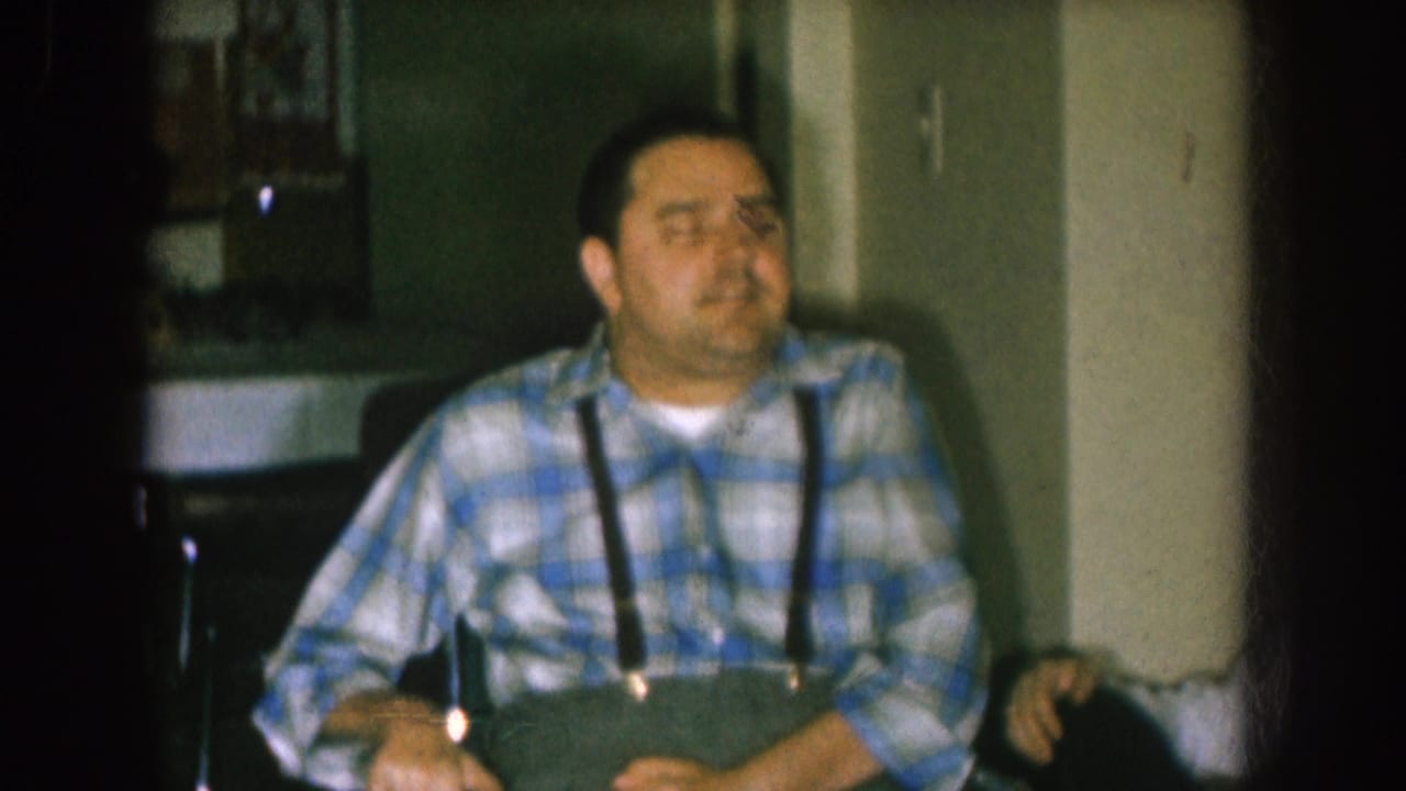 In 1957 Nebraska home-movie footage, a man in a blue plaid shirt and suspenders sits in a chair or wheelchair inside a dim home interior.