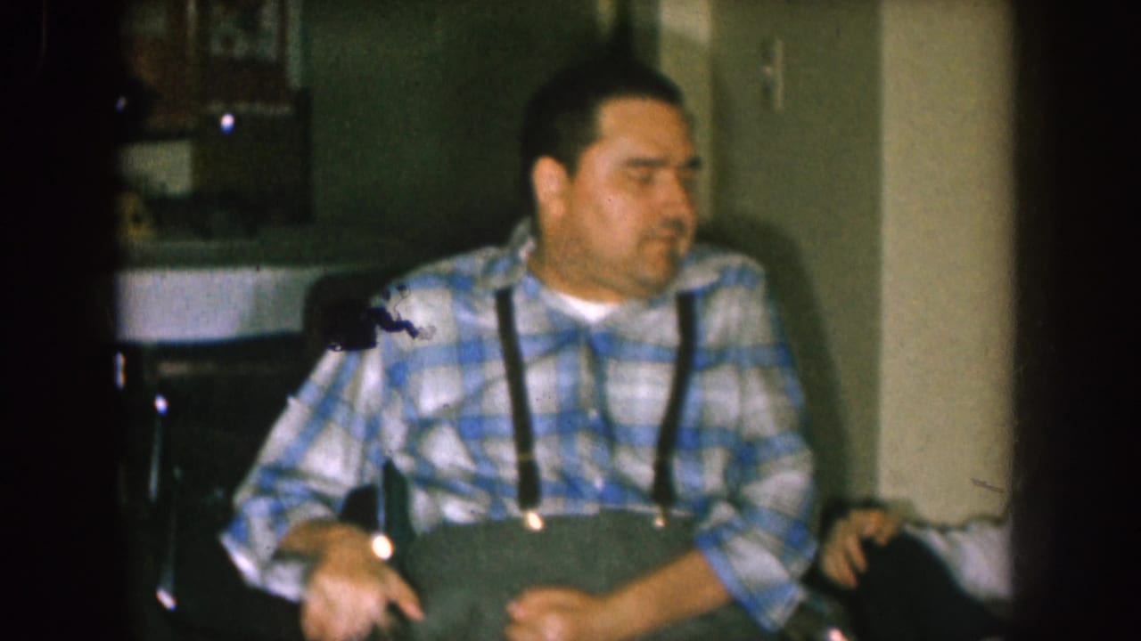 Seated man in plaid turns to one side in a dim interior.