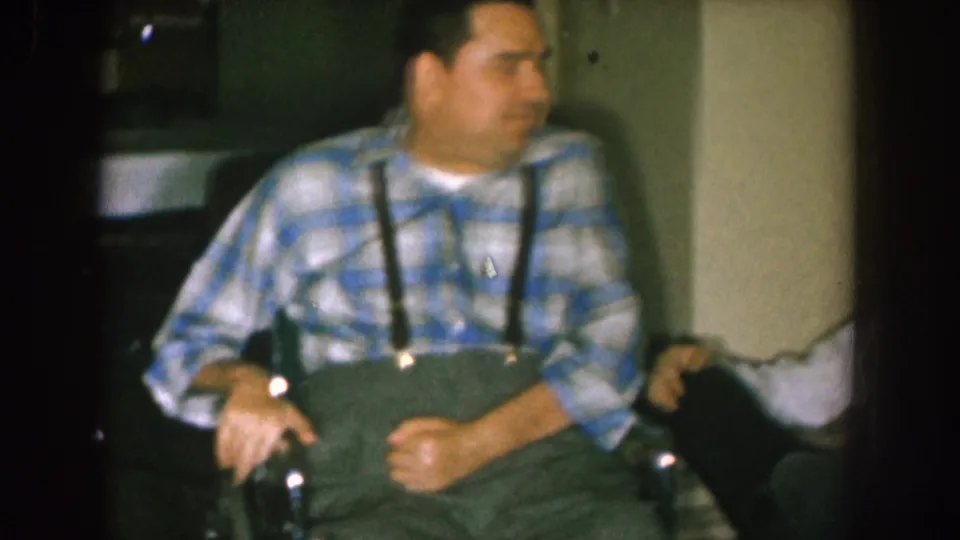 A man in a plaid shirt sits in a chair or wheelchair, with another person partly visible beside him in the 1957 Nebraska home interior.