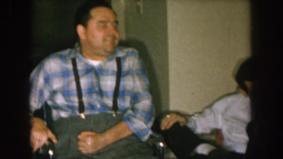 The seated man in a blue plaid shirt appears to smile near a partially visible person in a white shirt inside the 1957 Nebraska home setting.