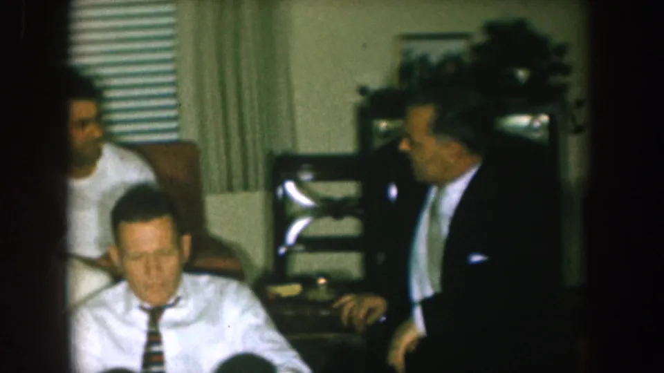 1957 Nebraska home-movie frame showing a suited man near seated family members in a private living room.