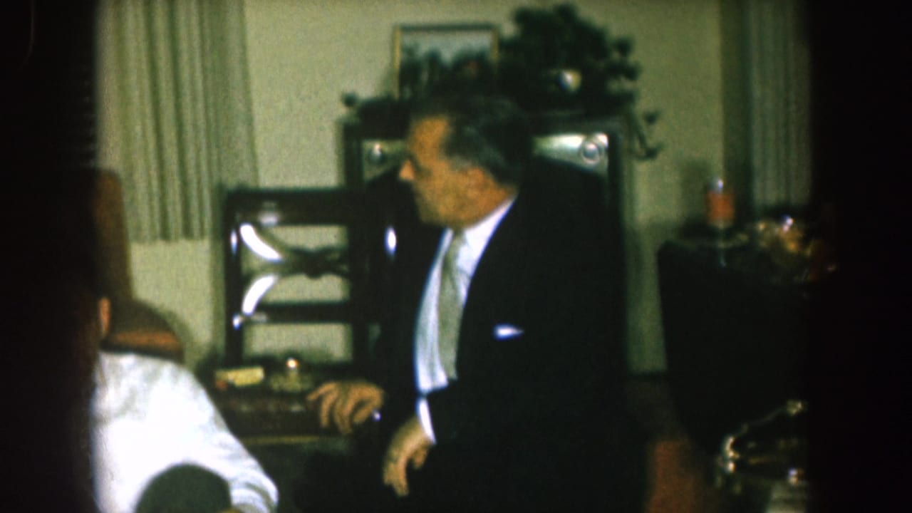 1957 Nebraska home-movie frame showing a suited man standing beside seated adults in a formal living room gathering.