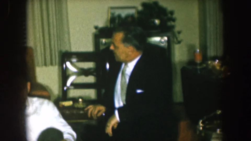 1957 Nebraska home-movie frame showing a suited man standing beside seated adults in a formal living room gathering.