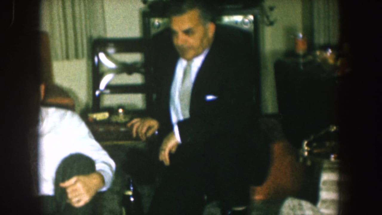 Man in a dark suit gestures beside seated adults and living-room furniture.