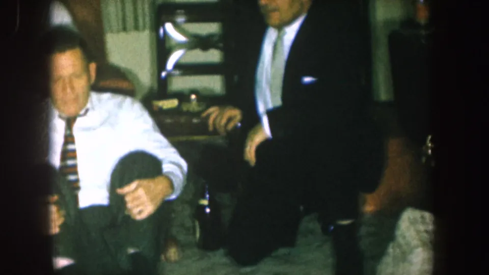 1957 Nebraska home-movie frame showing a seated man in a white shirt and striped tie next to a standing suited guest.