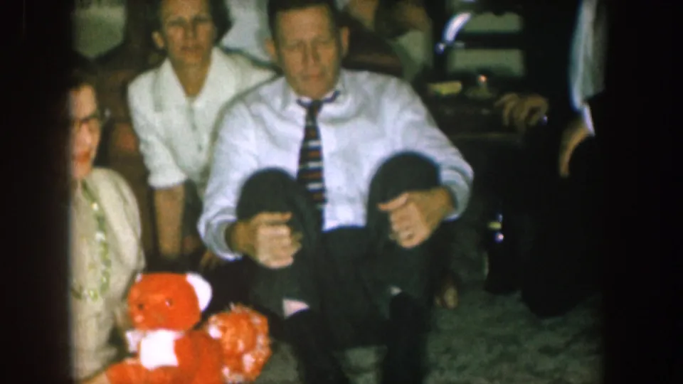 1957 Nebraska frame showing adults seated closely in a living room, with formal clothing and a red floral arrangement visible.