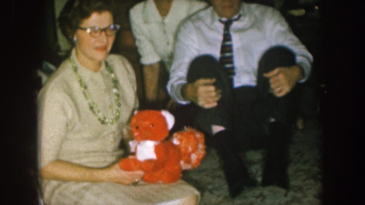 In a 1957 Nebraska home-movie frame, a woman in a cream dress holds a red teddy bear as men sit close beside her.