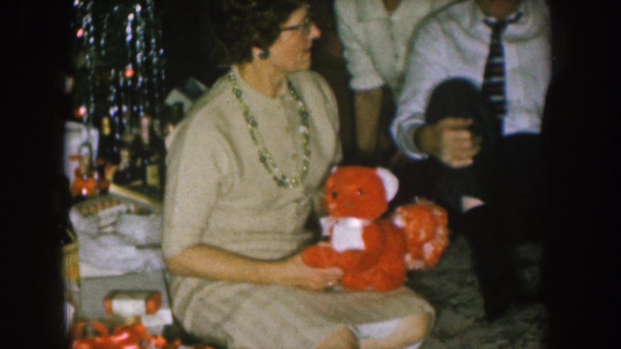 Woman holding a red teddy bear turns toward a seated man in a striped tie.
