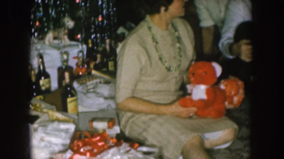 Wrapped gifts and tinsel appear beside the seated woman as she holds the red teddy bear in the 1957 Nebraska interior.