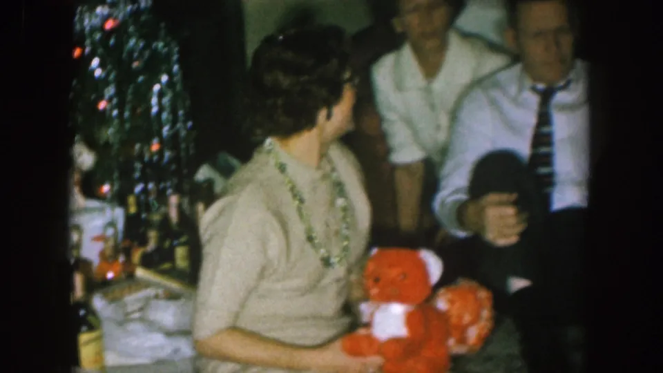 The woman looks back toward seated men while holding the red teddy bear in a private Nebraska holiday interior.