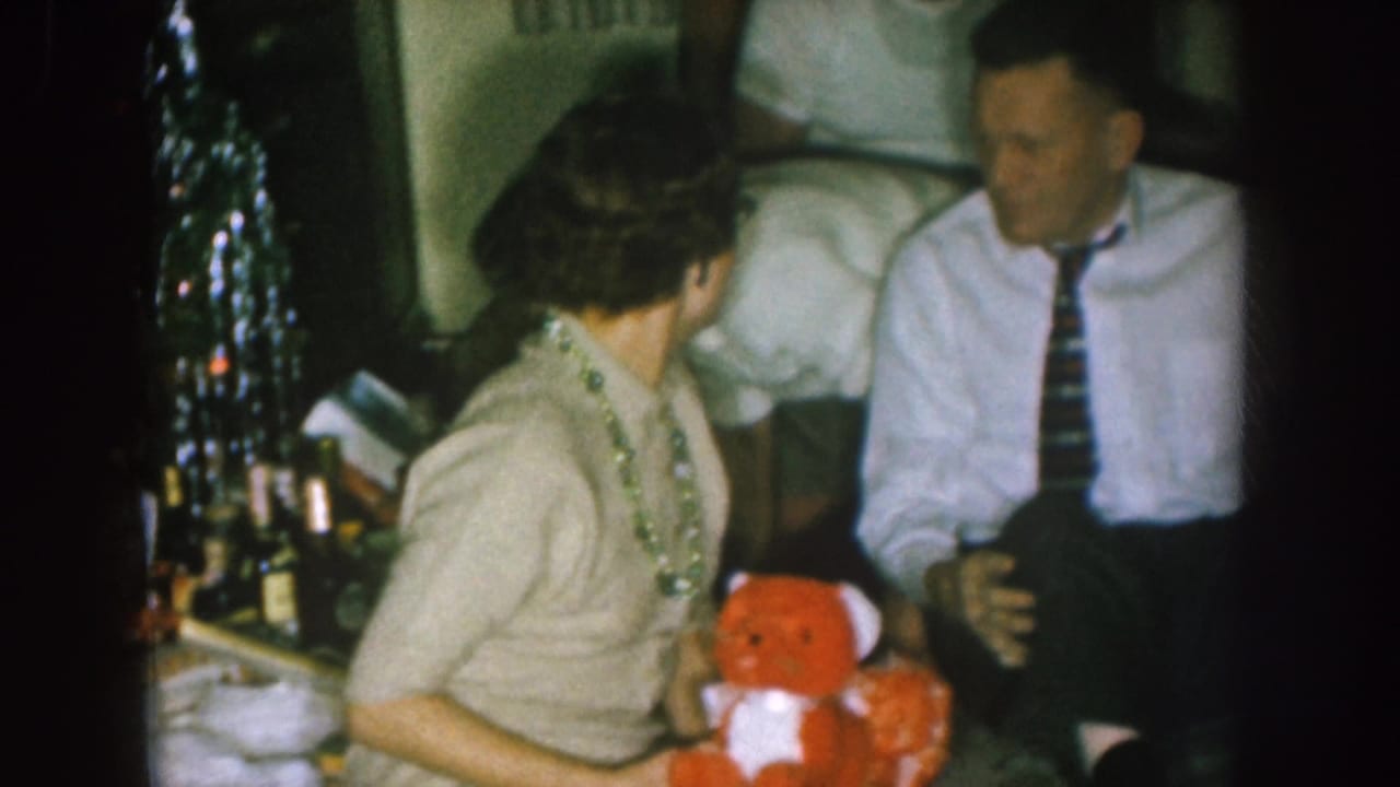 Adults seated near a Christmas tree, with a woman holding a red teddy bear beside a man in a white shirt and striped tie.