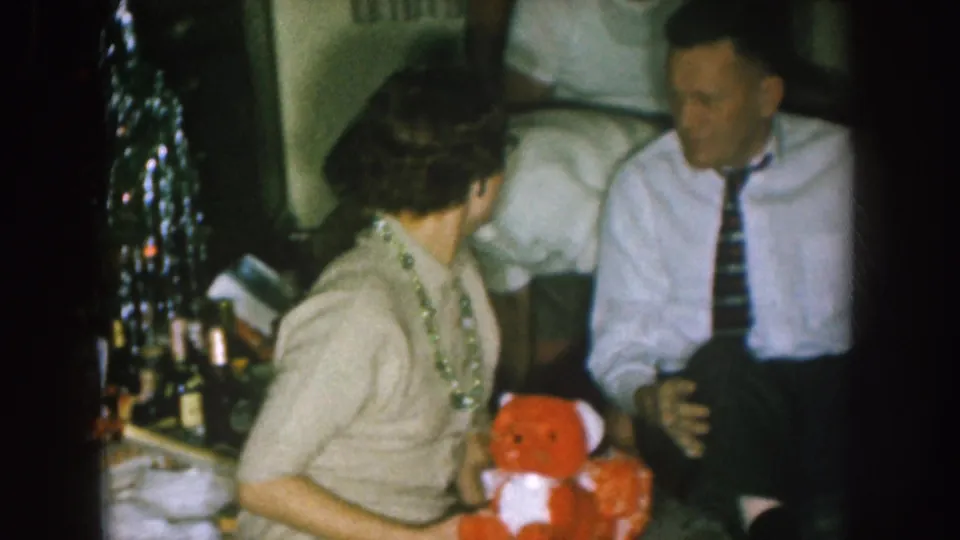 1957 Nebraska home-movie frame showing adults gathered indoors near a decorated Christmas tree as a woman holds a red teddy bear.