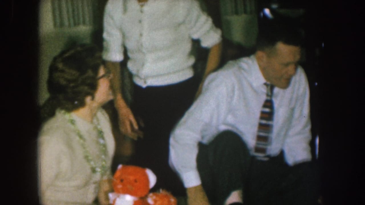 Woman seated with a red teddy bear while a man in a white shirt sits beside her and another adult stands behind them.