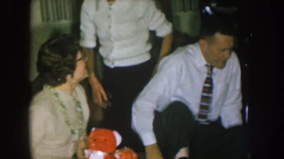 1957 Nebraska home-movie frame of adults in a private room near holiday decorations, with a red teddy bear visible in the foreground.