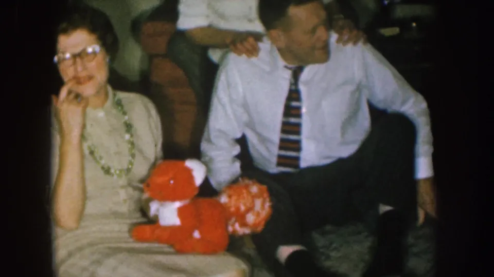 1957 Nebraska home-movie frame showing a woman with glasses and beaded necklace seated with a red teddy bear during a family gathering.