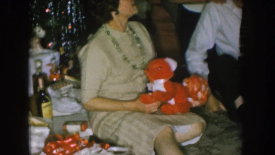 1957 Nebraska home-movie frame of a woman seated near a Christmas tree with a red teddy bear and holiday items in a private interior.