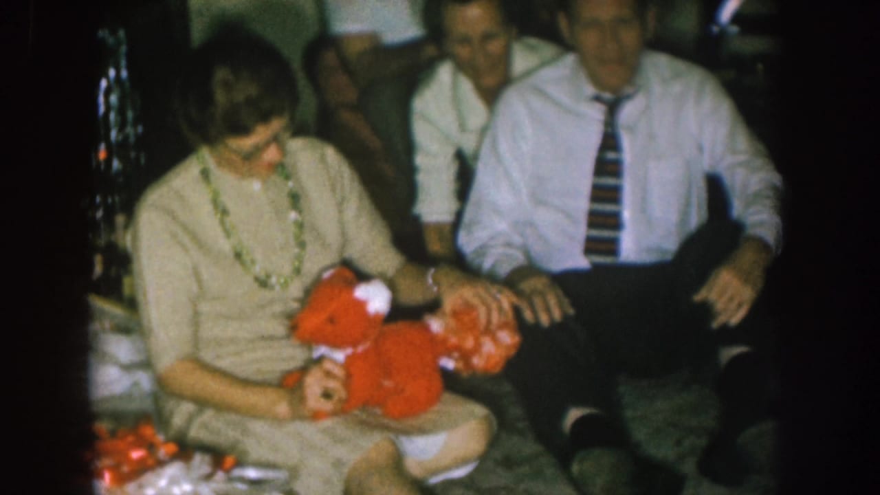 Woman in beige dress holds a red stuffed toy beside a seated man in a striped tie.