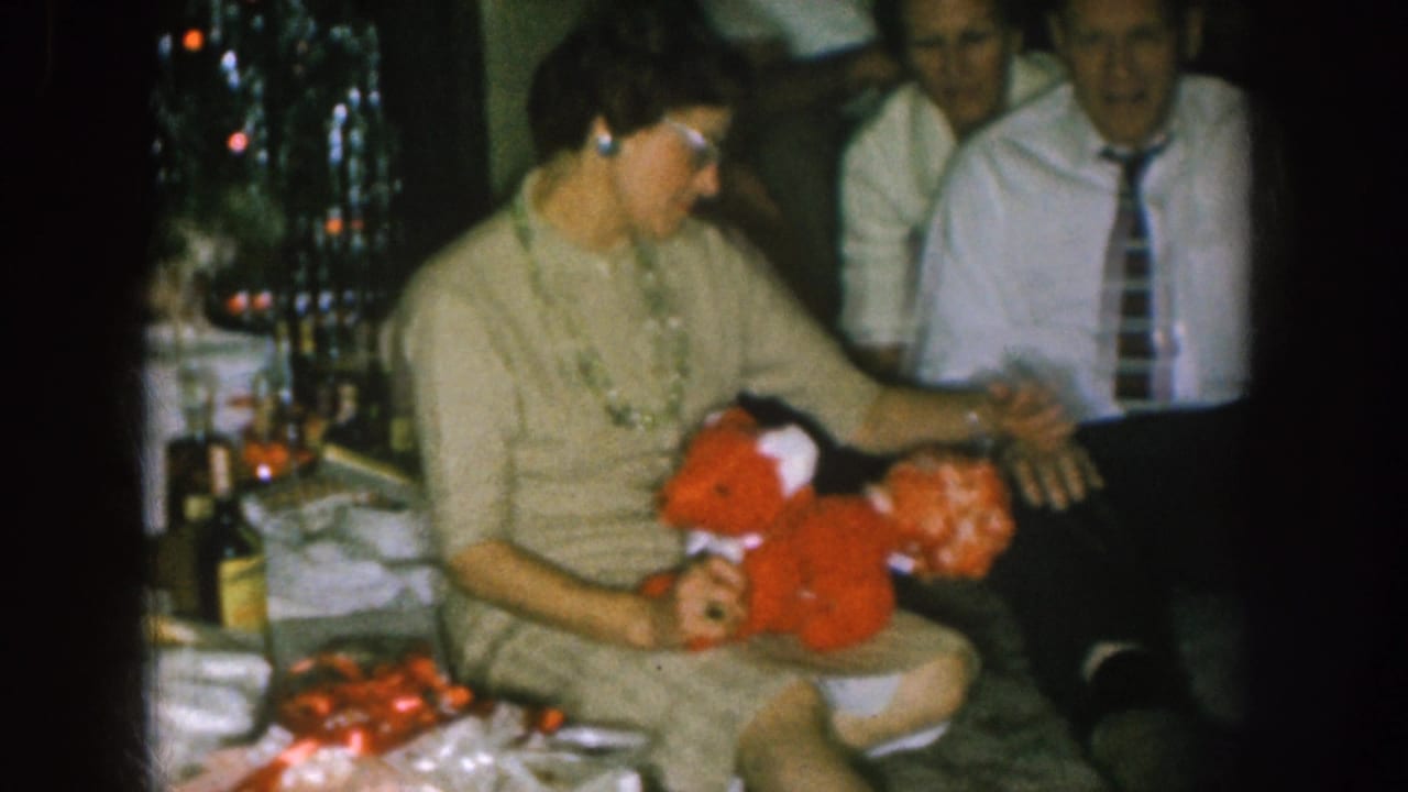 Woman turns toward a man while holding the red stuffed toy in a dim room.