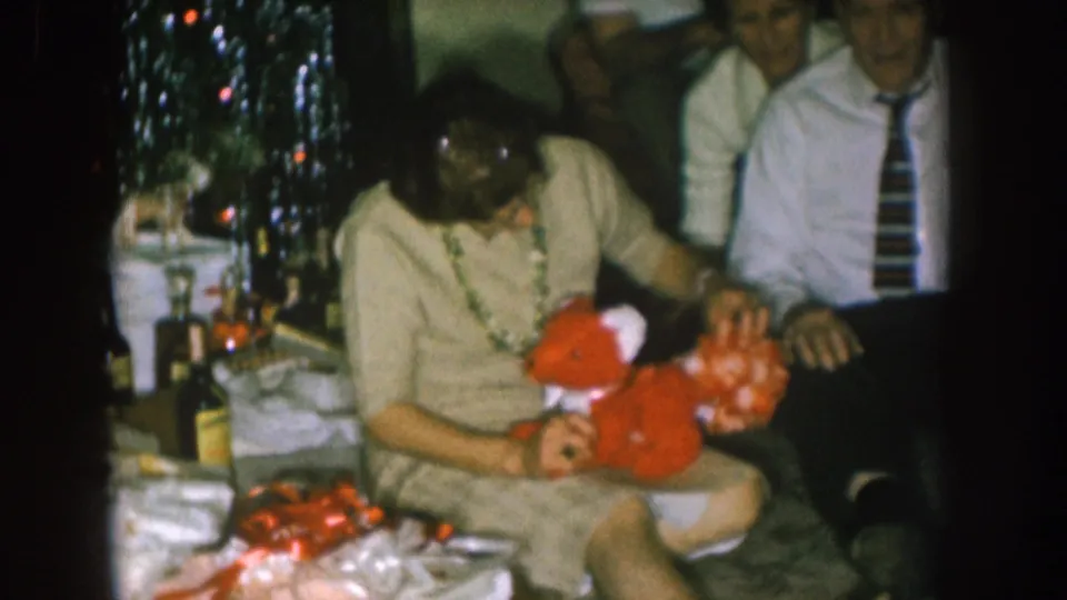 Indoor 1957 Nebraska home-movie frame showing a woman examining a red stuffed fox toy with adult guests seated nearby.