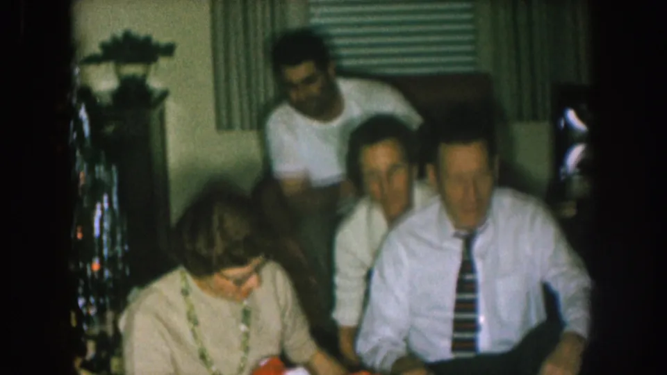 1957 Nebraska home-movie frame with seated adults gathered in a private interior near holiday decorations and wrapped gifts.