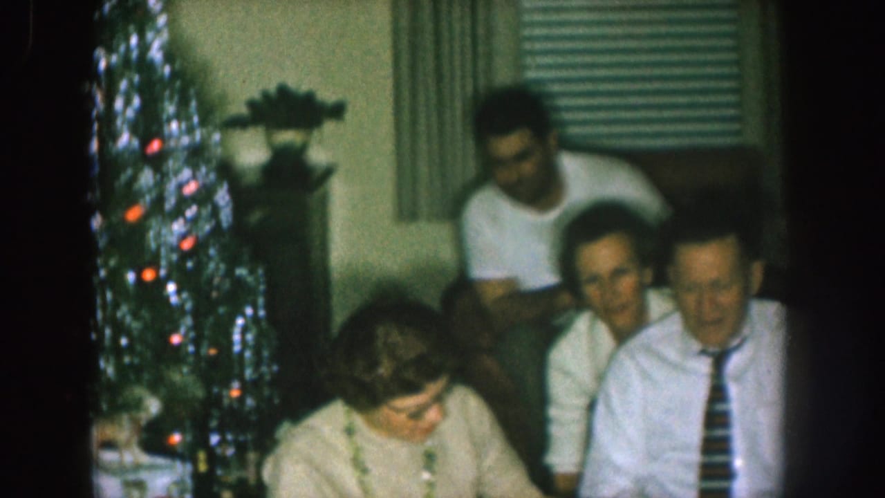 1957 Nebraska home-movie frame showing adults gathered in a living room with a decorated Christmas tree, curtains, and venetian blinds.