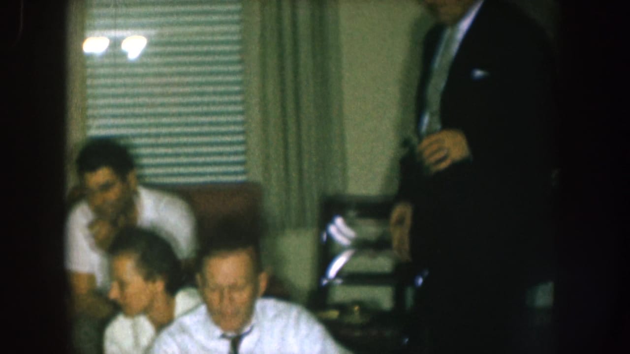 Several men sit and stand in a living room during a gathering.