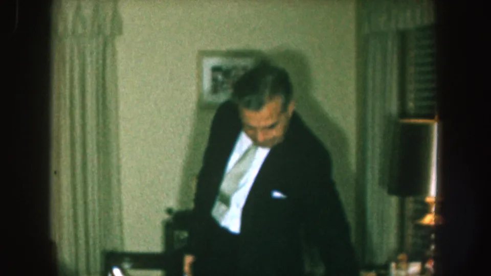1957 Nebraska home-movie frame showing a man in a dark suit and light tie standing beside living-room furniture and a table lamp.