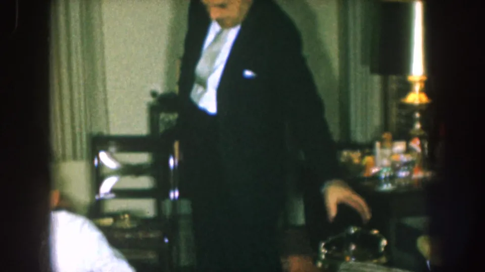 1957 Nebraska frame showing a man in a dark suit moving across a private living room with chairs, curtains, lamps, and side tables.