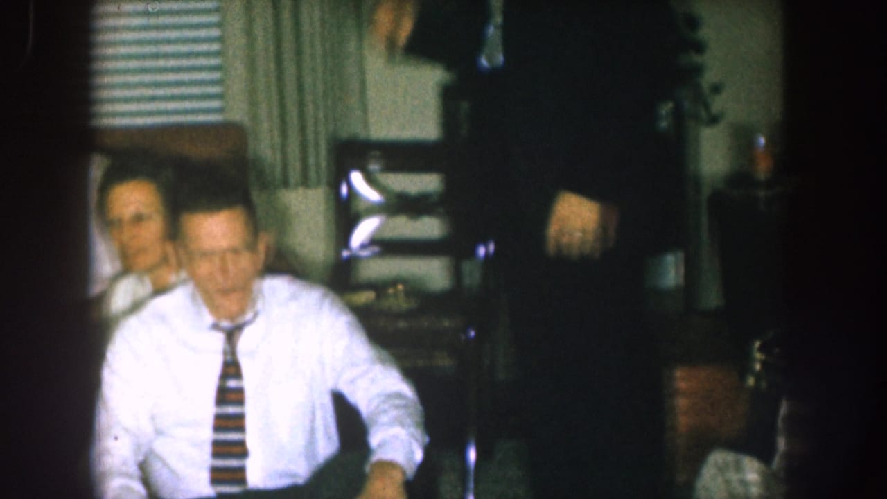 Man in white shirt and striped tie seated in a living room with another adult behind him.