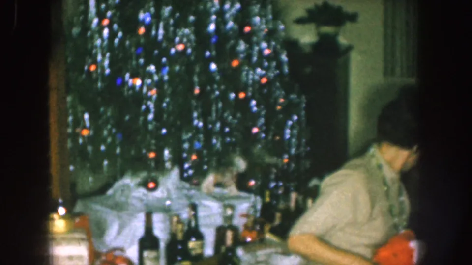 1957 Nebraska home-movie frame showing a lit Christmas tree and adults near a table in a private holiday interior.