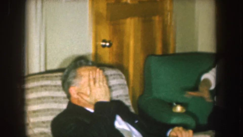 1957 Nebraska home-movie frame showing a suited man covering his face in a living room beside a green armchair.