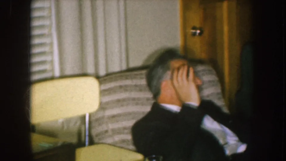 1957 Nebraska home-movie frame showing a side view of a seated man covering his face in a private living room.