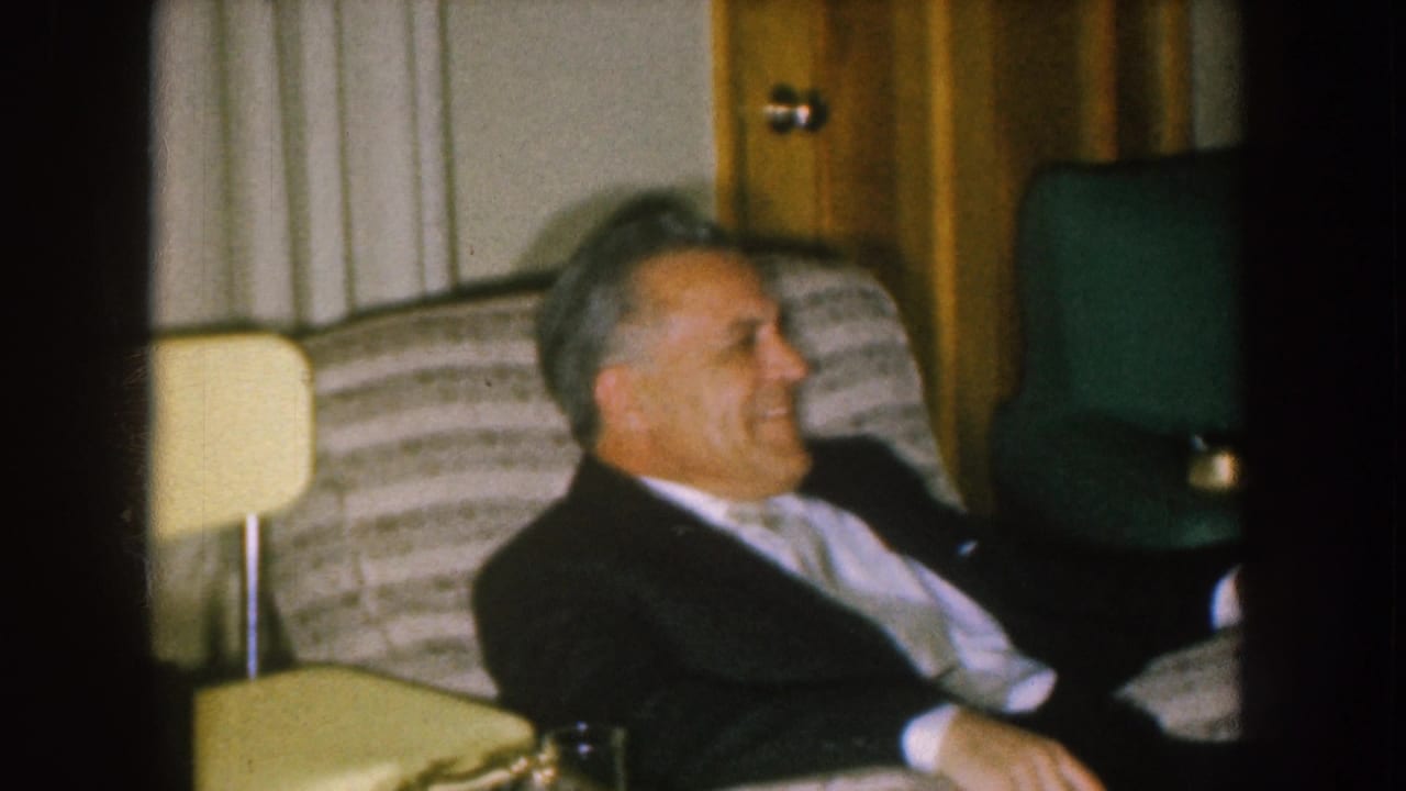 Man in a dark suit seated in a striped armchair beside a small lamp.