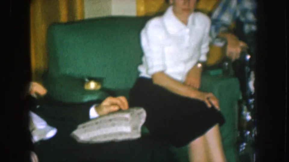 A woman in a white blouse sits near a green upholstered chair as other adults are partly visible in the Nebraska home interior.