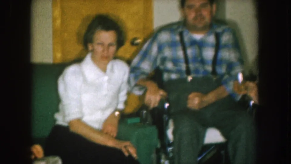 A woman sits beside a man in a plaid shirt and suspenders in a 1957 Nebraska private living room scene.