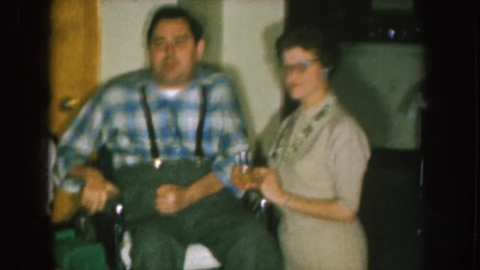 A softly focused 8mm frame shows the wheelchair, the man's plaid shirt and suspenders, and a woman in a light dress indoors.