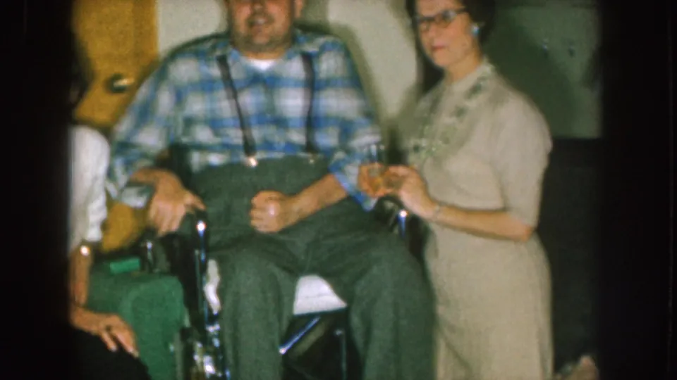 A 1957 Nebraska home-movie frame shows the seated man, the woman in a light dress, and part of another person at the edge of the frame.