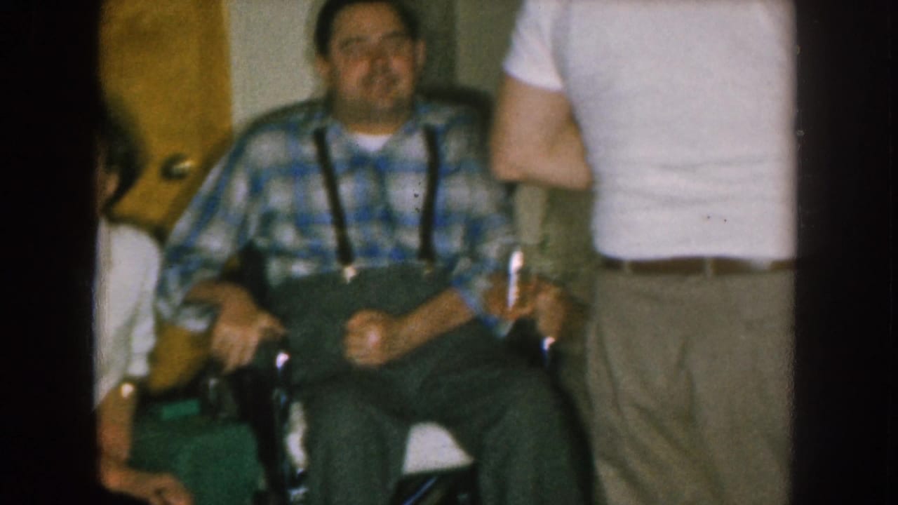 1957 Nebraska home-movie frame showing adults in a kitchen, with a seated man in plaid suspenders and partial figures nearby.