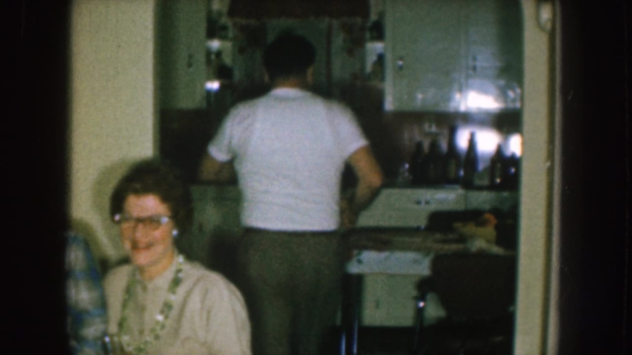 Woman in glasses sits near a kitchen doorway while a man in a white T-shirt faces the counter.