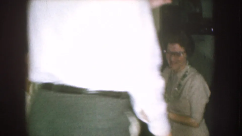 A close passerby creates a bright blur across the 1957 Nebraska home-movie frame, with a seated woman still visible in the kitchen.