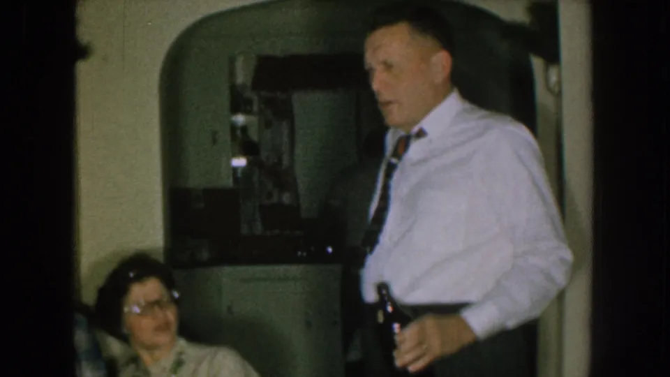 1957 Nebraska home-movie frame showing a man holding a bottle near a seated woman in glasses inside a private kitchen.