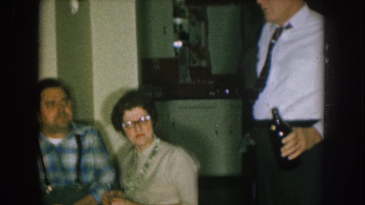 Adults seated and standing in a dim room, including a man in plaid and a woman with glasses.