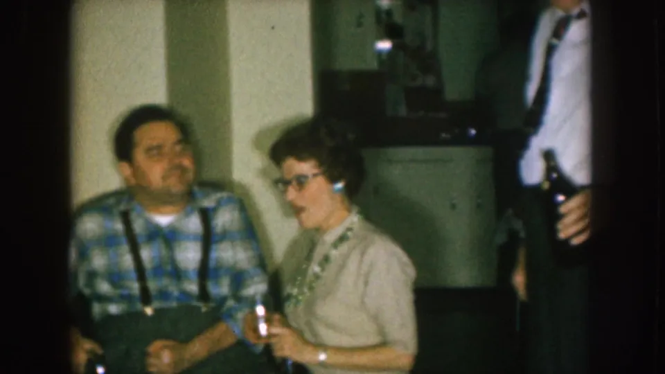 1957 Nebraska domestic gathering with a seated man in plaid and suspenders, a woman with glasses holding a glass, and a standing adult with a bottle.