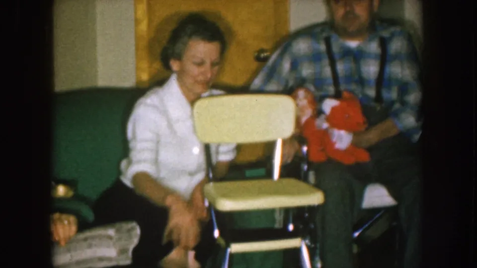 1957 Nebraska home-movie frame with a woman beside a folding highchair and a seated man in plaid holding a bright red object in a private room.